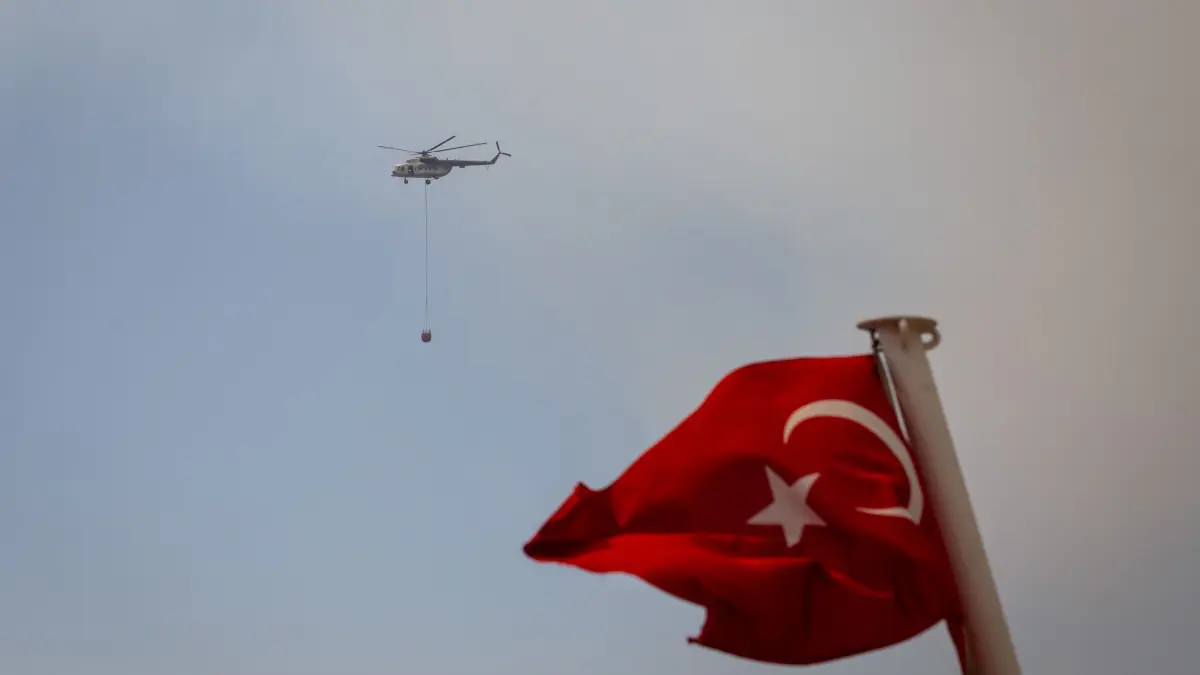 Waldbrände bei Izmir: 16.08.2024, Türkei, Izmir: Ein Löschhubschrauber fliegt hinter einer türkischen Fahne über die Bucht. Waldbrände bedrohen die westtürkische Küstenmetropole Izmir. Einsatzkräfte bekämpften das Feuer in der Nacht zum Freitag vom Boden und aus der Luft, wie die staatliche türkische Nachrichtenagentur Anadolu berichtete. Foto: Tolga Ildun/ZUMA Press Wire/dpa +++ dpa-Bildfunk +++