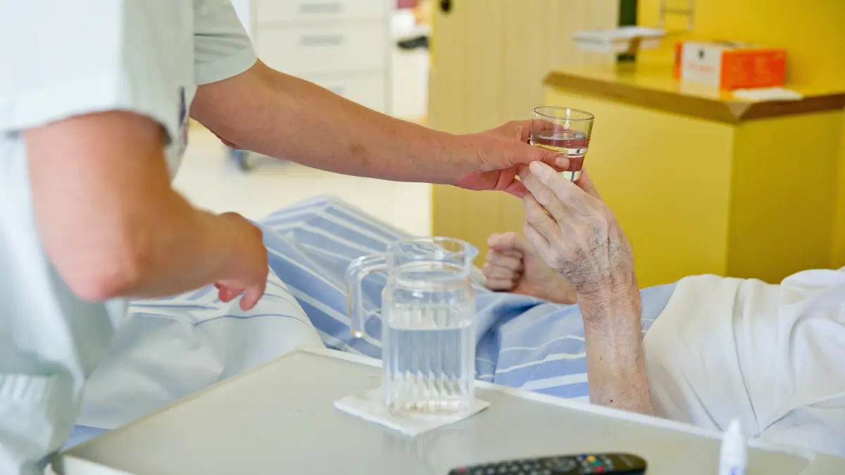 Eine Krankenpflegerin reicht einem Patienten in einem Klinikum ein Glas Wasser (Symbolbild). Gerade in Krankenhäusern leiden die Menschen unter der Hitze. Patientenzimmer sind oftmals nicht klimtisiert.