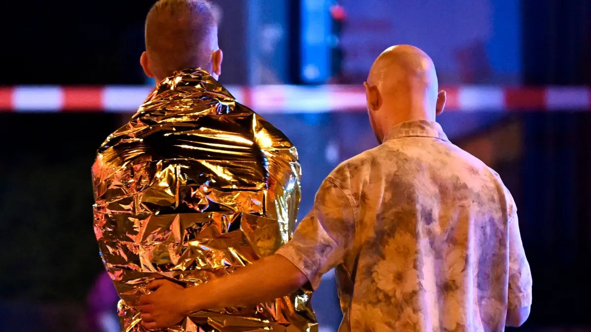 GERMANY-LGBTQ+-PARADE-CSD-POLICE: A man comforts another one who is wrapped in a security blanket near the scene where one person was killed and around 15 injured after a car is believed to have hit the crowd on the outskirts of the Christopher Street Day (CSD) parade in Berlin, on July 25, 2026. "We believe that a vehicle entered the Tiergarten park and hit several people and injured them," police posted on social media. The injured were being treated by the emergency services and they were actively seeking the people responsible, they added. (Photo by RALF HIRSCHBERGER / AFP)