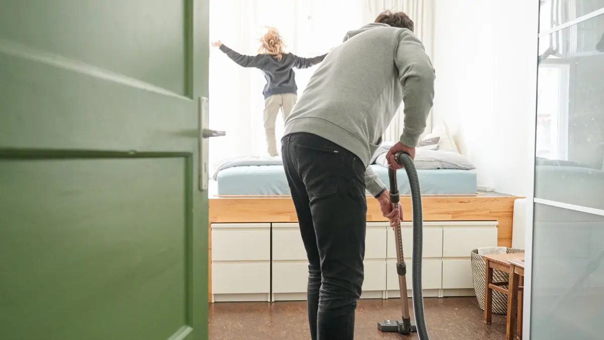 Ein Mann saugt Staub während ein Kind auf dem Bett hüpft: ILLUSTRATION - 12.02.2022, Berlin: Kinderbetreuung, Haushalt und Beruf lassen sich nicht immer leicht unter einen Hut zu bringen. (zu dpa: «Umfrage: Familie und Beruf noch immer schwer zu vereinbaren») Foto: Annette Riedl/dpa/dpa-tmn +++ dpa-Bildfunk +++