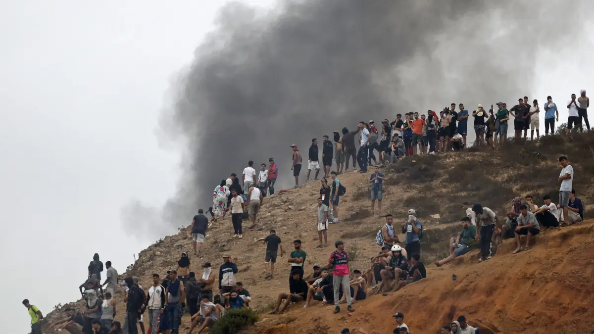 MOROCCO-SPAIN-MIGRATION: People gather on a hilltop as smoke billows from the site of clashes near Fnideq, on the Morocco-Spain border, on July 31, 2026. Thousands of migrants crossed overnight from Morocco into Spain's north Africa's Ceuta enclave, a police source said on July 31, adding that it was impossible to estimate how many people in total. A total of 18 people died while attempting to reach the Spanish enclave, the source said. (Photo by Abdel Majid BZIOUAT / AFP)