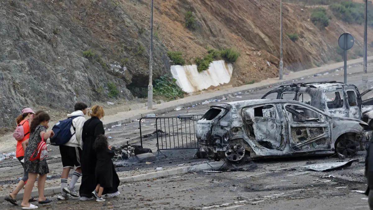 MOROCCO-SPAIN-MIGRATION: People walk past burnt vehicles at the site of clashes near Fnideq, on the Morocco-Spain border, on July 31, 2026. Thousands of migrants crossed overnight from Morocco into Spain's north Africa's Ceuta enclave, a police source said on July 31, adding that it was impossible to estimate how many people in total. A total of 18 people died while attempting to reach the Spanish enclave, the source said. (Photo by Abdel Majid BZIOUAT / AFP)