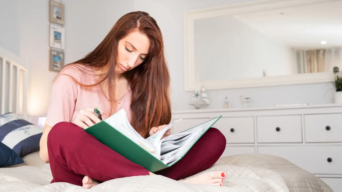 Junge Frau sitzt mit einem Aktenordner auf einem Bett: ILLUSTRATION - Zum Ausbildungsstart sollten sich Berufseinsteiger auch mit dem Thema Versicherungen auseinandersetzen. (zu dpa: «Versicherungen und Finanzen: Was Azubis jetzt wissen sollten») Foto: Franziska Gabbert/dpa-tmn - Honorarfrei nur für Bezieher des dpa-Themendienstes +++ dpa-Themendienst +++