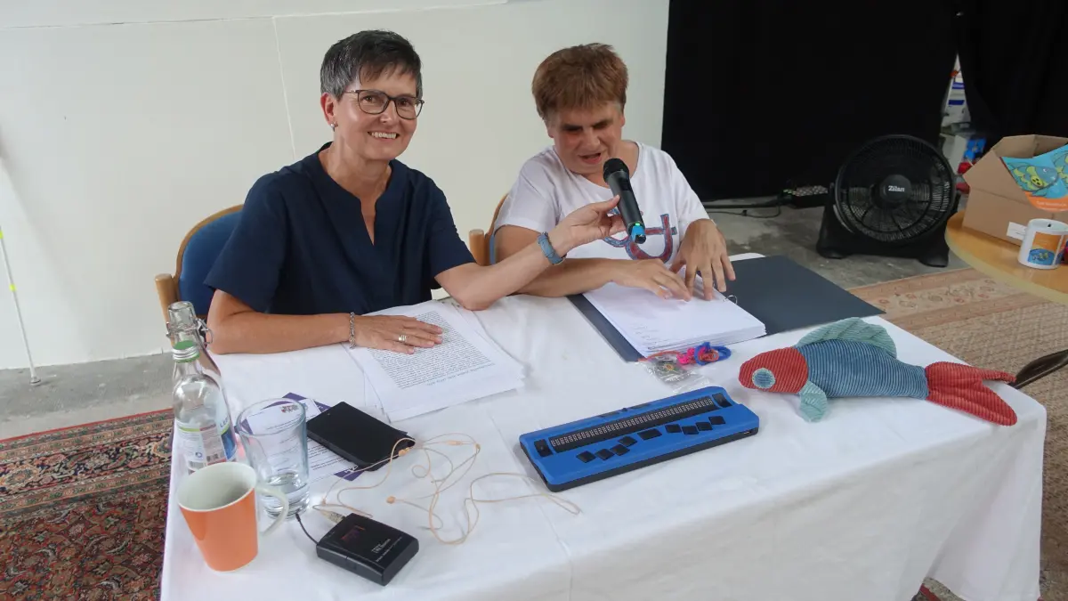 Simone Moosmann (links) liest als sehende Assistenz in Schwarzschrift mit, während Sabine Ludi die ausgewählten Kapitel in Brailleschrift ertastet und vorliest. Mit dabei: Ludis treuer Begleiter „Sehnix“.