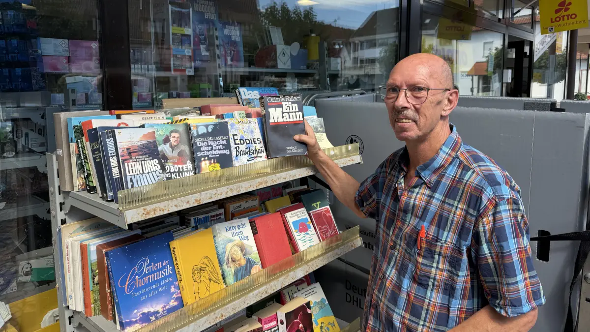 Hubert Schuhmacher steht neben seinem Bücherregal – wo man kostenfrei Bücher mitnehmen und ablegen kann.