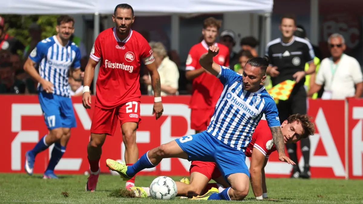 Bahlinger SC - 1. FC Magdeburg: 23.08.2026, Baden-Württemberg, Bahlingen am Kaiserstuhl: Fußball, Männer: DFB-Pokal, Bahlinger SC - 1. FC Magdeburg, 1. Runde, im Kaiserstuhlstadion, Barış Atik (2.v.r., 1. FC Magdeburg) und Holger Bux (Bahlinger SC) kämpfen um den Ball. Foto: Philipp von Ditfurth/dpa - WICHTIGER HINWEIS: Gemäß den Vorgaben der Bundesliga bzw. des DFB Deutscher Fußball-Bund ist es untersagt, in dem Stadion und/oder vom Spiel angefertigte Fotoaufnahmen in Form von Sequenzbildern und/oder videoähnlichen Fotostrecken zu verwerten bzw. verwerten zu lassen. +++ dpa-Bildfunk +++