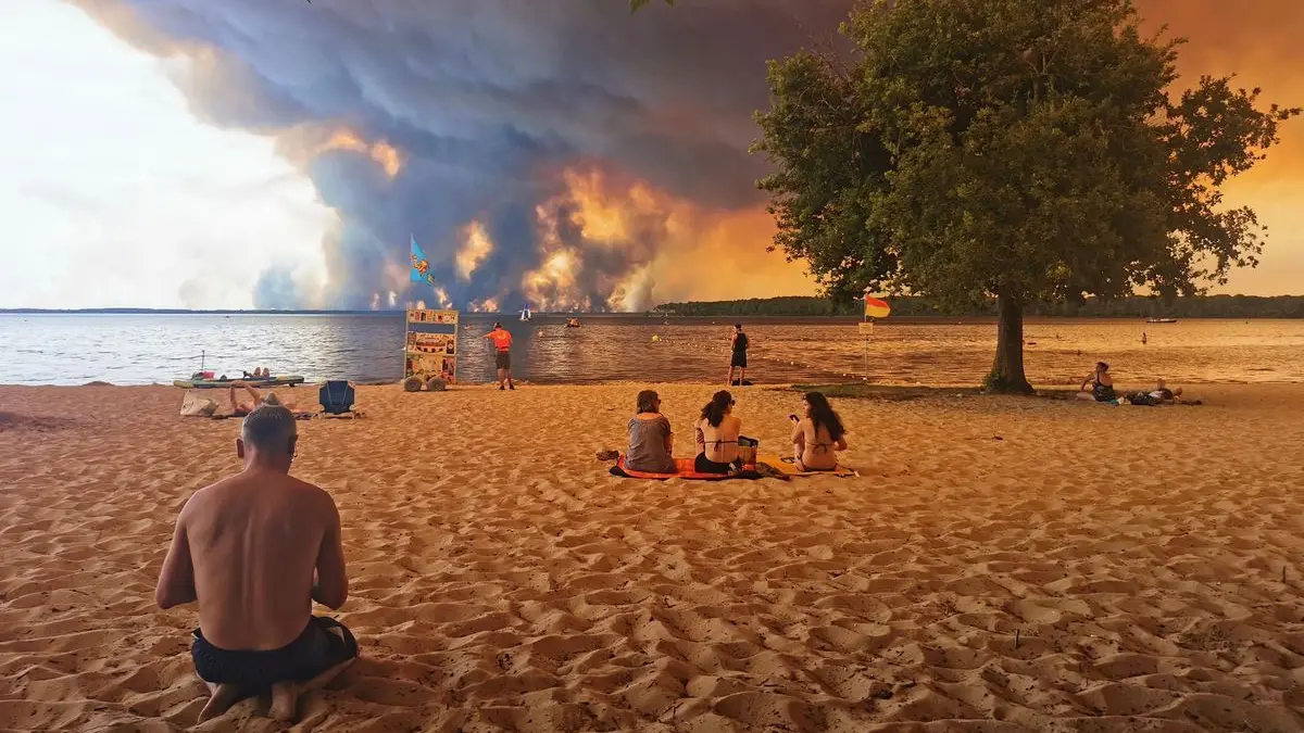 Strandbesucher sitzen im Juli 2026 am Strand von Moutchic in Lacanau im Südwesten Frankreichs, während Rauchwolken von einem Waldbrand in den Himmel aufsteigen.