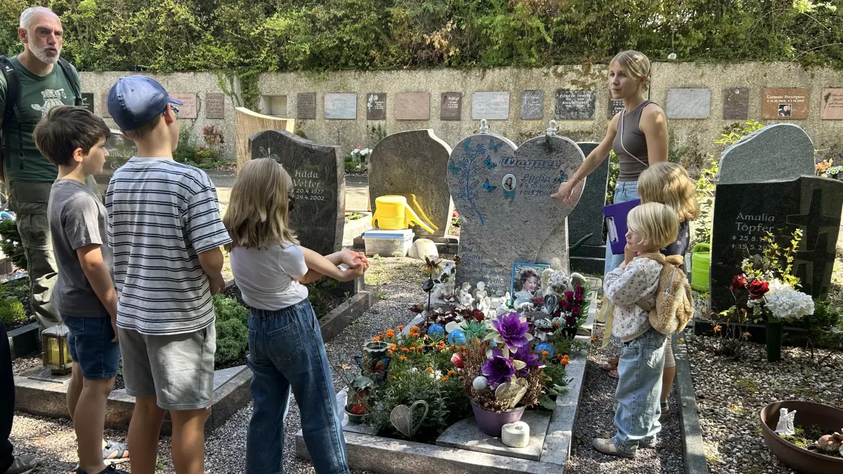 Achim Wenzel-Teuber vom Netzwerk Trauer und Friedhofsleiterin Sarah Bonomo zeigen Kindern das Grab einer jungen Frau.