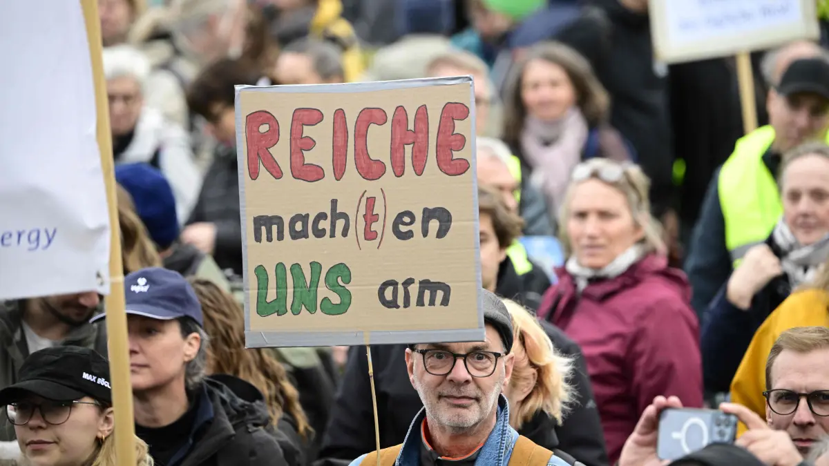 GERMANY-POLITICS-GOVERNMENT-ENERGY-DEMO: A protester holds up a placard reading "The Rich Make Us Poor / (German Economy and Energy Minister Katherina) Reiche Makes Us Poor" during a demonstration by Germany's wind energy sector against Economy Ministry reforms in front of the Reichstag building in Berlin on October 9, 2026. (Photo by Tobias SCHWARZ / AFP)