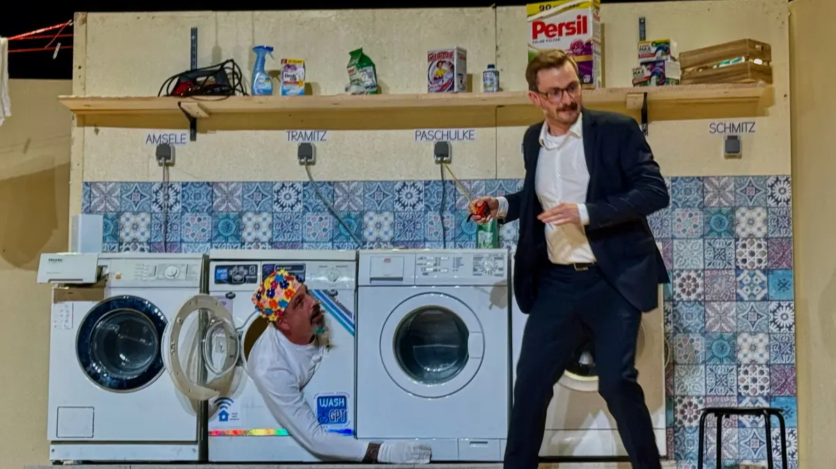 Die KI wird handgreiflich: WashGPT (Matthias Veith) greift aus der Trommel nach Marco Meininger (Moritz Ragg), der die Waschküche in einen Solarbetrieb umbauen will.